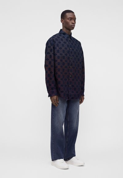 Homme portant une chemise sombre à motifs avec boutons, un jean bleu ample et des baskets blanches, debout devant un fond uni.