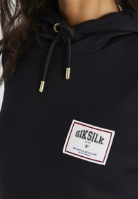 Černá mikina s kapucí a šňůrkami, vybavená zlatými detaily a bílou značkovou nášivkou s červenými akcenty. Měkký, elastický materiál a slim fit střih.