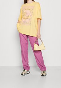 Camiseta gráfica amarilla de gran tamaño con estampado de palmeras, combinada con pantalones de chándal rosas. Los accesorios incluyen un pequeño bolso amarillo y zapatillas deportivas.