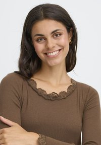 Langærmet brun bluse med ribbet tekstur, der har en bølget kant af blonder ved halsudskæringen og manchetterne, som fremhæver det feminine design.