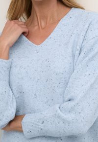 Jasnoniebieski sweter o fakturowanej strukturze, z dekoltem w serek, ściągaczami na rękawach oraz rozproszonymi czarnymi nitkami w całej tkaninie. Wygodne dopasowanie i miękki materiał.