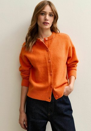 Cárdigan naranja hecho de un material suave, con cuello redondo, botones frontales y puños acanalados. Combinado con jeans oscuros.