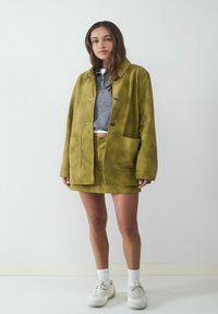 Olivgrüne Wildlederjacke mit zwei Fronttaschen, kombiniert mit einem grauen Cropped-Top und passendem Minirock über weißen Sneakers.