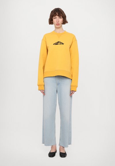 Gul genser med svart logo, vide lyseblå jeans med frynset hem og svarte flate sko. Enkelt, avslappet design.