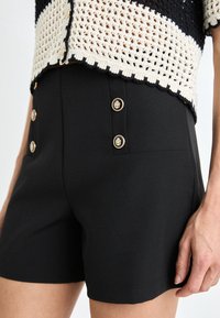 Czarne szorty z wysokim stanem, dwoma przednimi kieszeniami i złotymi guzikami jako dodatkowym akcentem. Gładka tekstura i dopasowany krój.