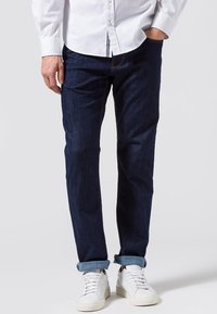 Jeans de mezclilla azul oscuro con un corte recto, con un dobladillo enrollado y combinados con zapatillas blancas. Camisa de botones de color neutro.