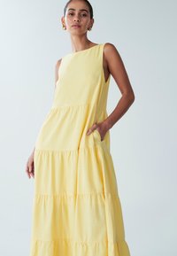 Gul ärmlös maxiklänning med flera volanglager, slät textur och sidofickor. Enkeldesign med fyrkantig urringning.