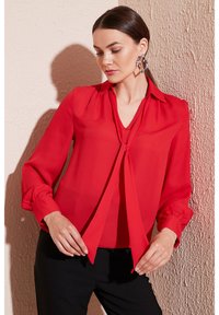 Red blouse made of lightweight fabric, featuring a neck tie, long sleeves with gathered cuffs, and a relaxed fit.
