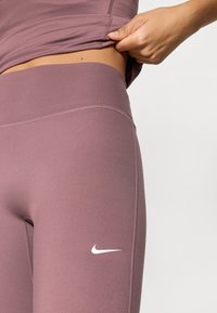 Κολάν Nike σε απόχρωση μοβ, κατασκευασμένα από ελαστικό ύφασμα. Διαθέτουν ψηλή μέση και μικρό λευκό λογότυπο στον αριστερό μηρό.
