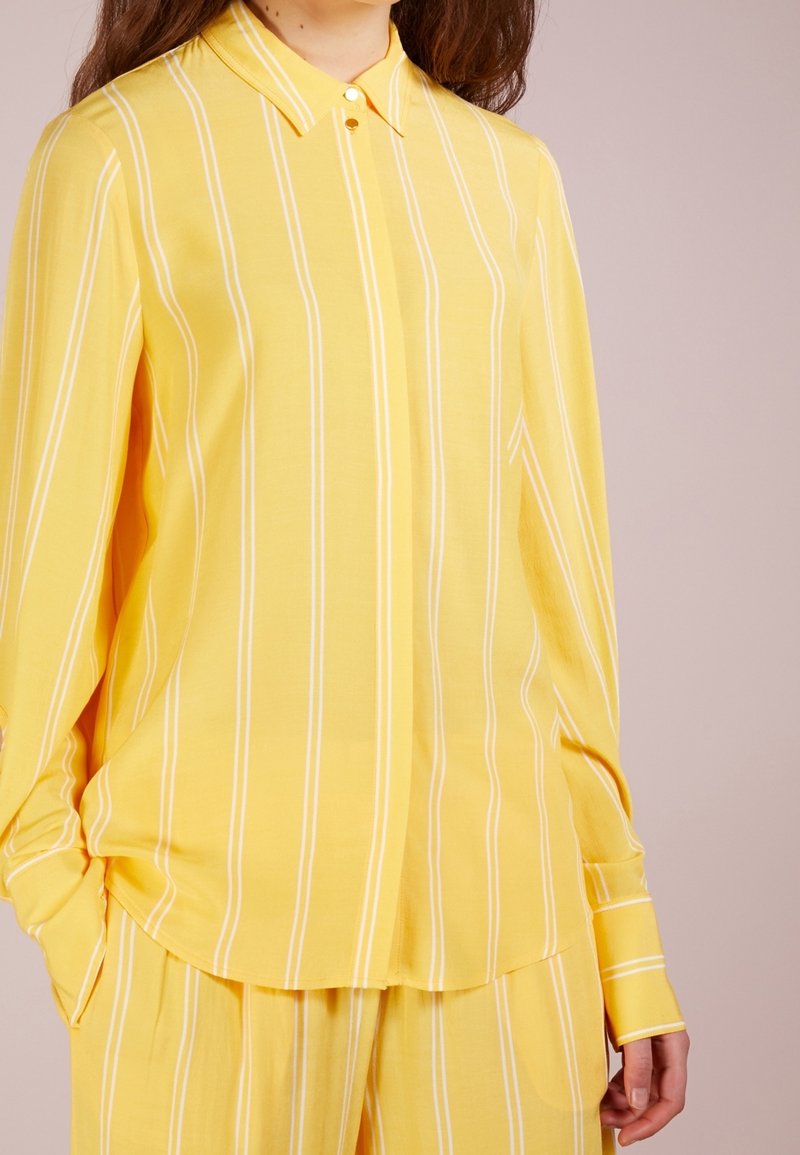 Camicia gialla a maniche lunghe con strisce verticali bianche, colletto con bottoni e vestibilità morbida; tessuto liscio con una leggera lucentezza.