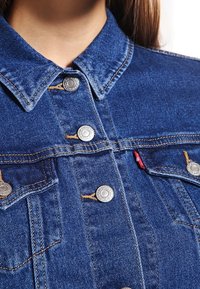 Lähikuva sinisestä denim-takista, jossa on metallinapit, keltasetomukset ja pieni punainen kangastunnus taskussa.