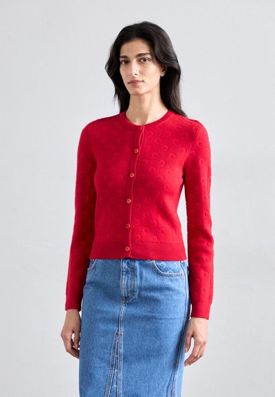 Cardigan texturat roșu cu decolteu rotund, mâneci lungi și închidere cu nasturi. Asortat cu o fustă din denim albastru cu o cusătură centrală.