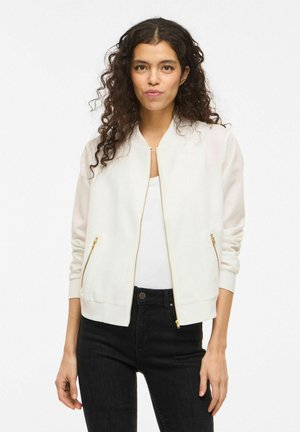 Kvinne med langt krøllete hår iført en hvit bomberjakke med glidelås over en hvit topp og svarte jeans, stående foran en ensfarget hvit bakgrunn.