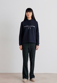 Sudadera azul marina con logo blanco "TOMMY HILFIGER" y cordón; combinada con pantalones negros y zapatos beige. Diseño simple y moderno.
