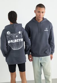 Felpa grigia con cappuccio e tasca frontale. Il design grafico sul retro presenta pianeti e la scritta "GALACTIC" in bianco. Tessuto morbido e texture.
