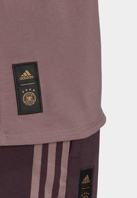 Odzież Adidas z krótkim rękawem w kolorze mauve oraz ciemnofioletowymi spodniami, obie z czarnymi metkami materiałowymi i złotymi akcentami logo.