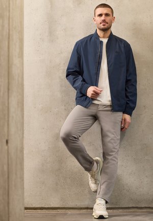 Mies, jolla on päällään tummansininen takki, valkoinen paita, beigehousut ja valkoiset tennarit, seisoo ristissä toinen jalka vasten betoniseinää.