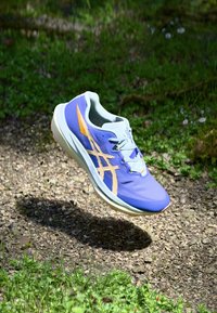 Könnyű, lila futócipő texturált felsőrésszel, kontrasztos narancssárga részletekkel és párnázott fehér középtalppal, amely kavics felett lebeg.