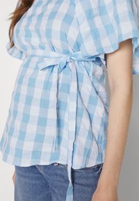 Top xadrez em azul claro e branco, feito de tecido texturizado, apresentando mangas curtas com babados e detalhe de cintura atada. Usado com denim.