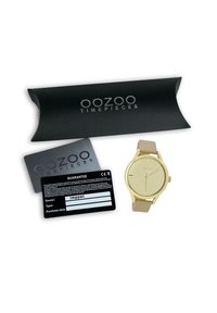 Montre dorée au design minimaliste, bracelet en silicone beige et cadran transparent. Accompagnée d'une carte de garantie noire et d'un emballage.
