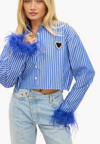 Chemise à rayures bleues et blanches, coupée, avec des poignets en plumes bleues et une broche en forme de cœur. Portée avec un jean en denim délavé.