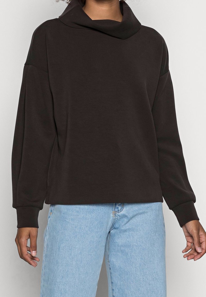 Personne portant un pull-over noir à manches longues avec un large col bénitier et un jean en denim bleu clair sur un fond uni.