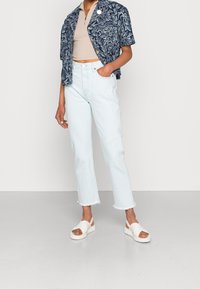 Marinblommig kortärmad jacka över en beige höghalstopp, kombinerad med ljusblå raka jeans och vita sandaler med tjock sula.