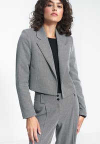 Żakiet w klasyczny wzór houndstooth w odcieniach szaro-białych, charakteryzujący się skróconym krojem, klapami i dwoma przednimi kieszeniami. W połączeniu z czarną bluzką.