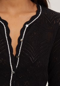 Cardigan noir en tricot avec un motif texturé, col en dentelle, bord blanc sur le devant et boutons ronds noirs.