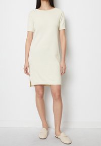 Abito lavorato a maglia di colore crema con maniche corte, scollo rotondo, spacchi laterali e orlo a coste. Abbinato a scarpe beige senza lacci.