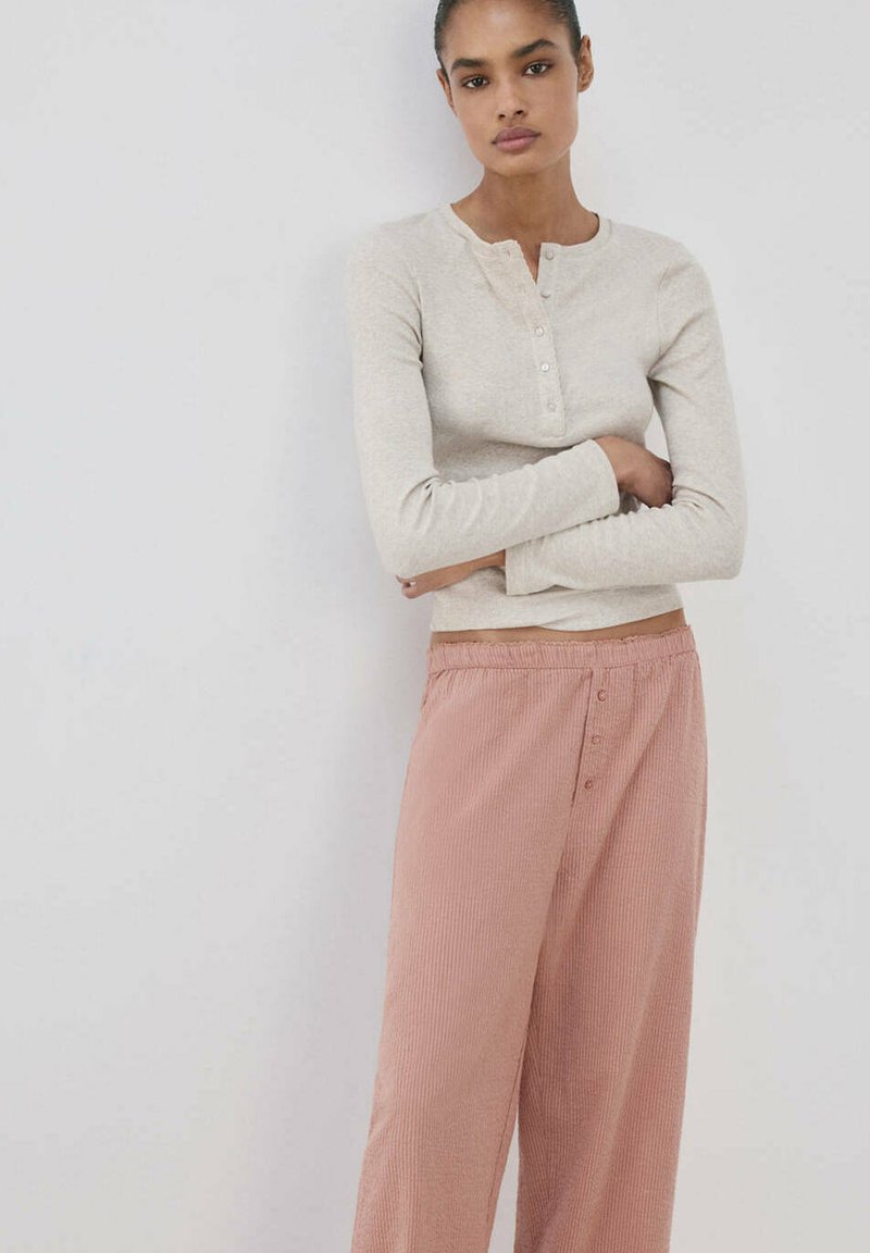 Jeune femme les bras croisés portant un haut à manches longues beige clair boutonné et un pantalon ample en velours côtelé rose sur un fond uni.