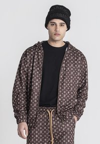 Chaqueta con cremallera de patrón marrón y detalles de logo en beige, combinada con camisa negra y joggers a juego que presentan un diseño similar. Acabada con un gorro negro.
