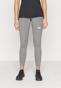 Γκρι κολάν από ελαστικό ύφασμα, με εφαρμοστή σχεδίαση και μικρό λευκό λογότυπο στο πάνω μέρος του μηρού. Συνδυάζεται με μαύρα αθλητικά παπούτσια.