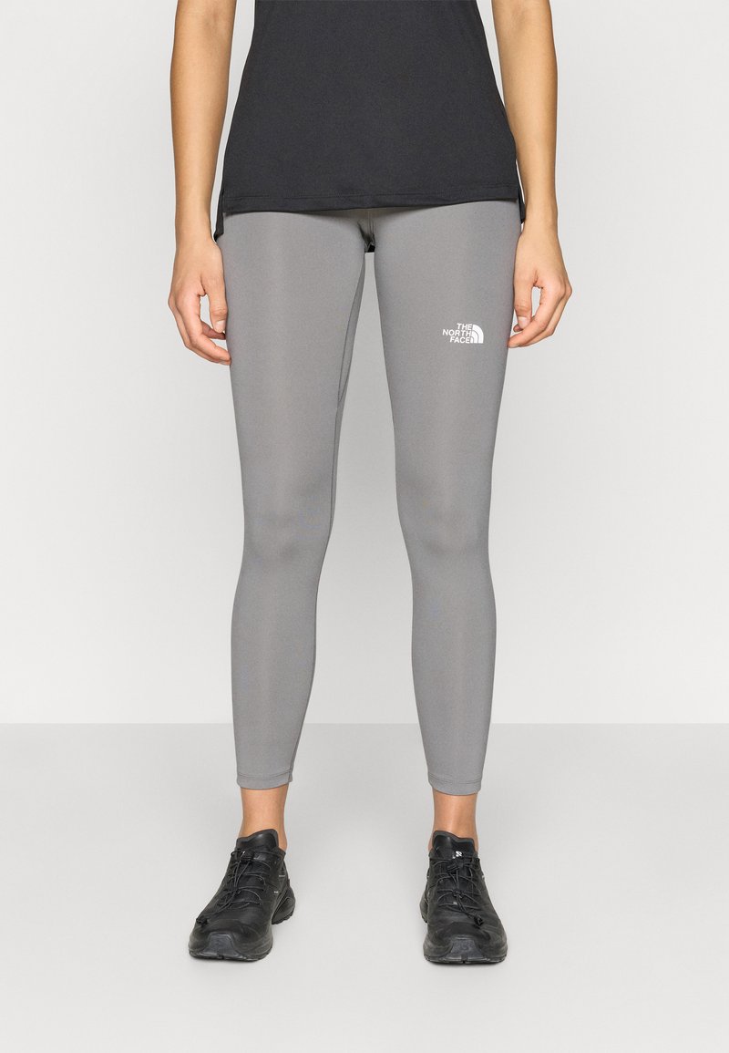 Γκρι κολάν από ελαστικό ύφασμα, με εφαρμοστή σχεδίαση και μικρό λευκό λογότυπο στο πάνω μέρος του μηρού. Συνδυάζεται με μαύρα αθλητικά παπούτσια.