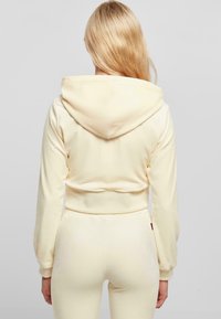 Crop hoodie en tissu beige clair et doux avec une capuche et des poignets ajustés, associé à un bas ajusté assorti. Texture lisse et détails minimalistes.