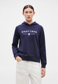 Námořnická mikina s kapucí a klokáním kapsou, s bílým nápisem "GANT1949" a štítovým logem. Měkká tkanina, žebrované manžety a lem. Minimalistický design.