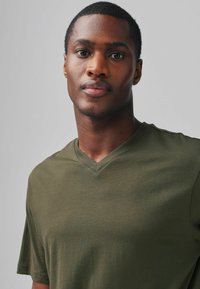 Jonge zwarte man met kort haar die een olijfgroen V-hals T-shirt draagt, recht vooruit kijkend met een neutrale uitdrukking tegen een effen grijze achtergrond.