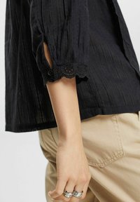 Schwarze Bluse mit Struktur und drei Viertelärmeln, ausgestattet mit Spitzenbesatz und dekorativem Knopf am Bund. Die Bluse wird mit beigen Hosen kombiniert.