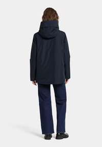 Marinblå vattentät jacka med huva, med en rak passform och slät textur, i kombination med mörk denimbyxor och kraftiga svarta skor.