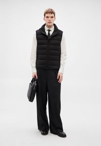 Μαύρο γιλέκο από φουσκωτό ύφασμα με ψηλό γιακά, φορεμένο πάνω από λευκό πουκάμισο και μαύρη γραβάτα. Μαύρο παντελόνι με φαρδιά μπατζάκια και λαμπερά μαύρα παπούτσια. Κρατάει μαύρη τσάντα χειρός.