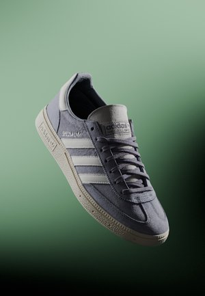 Baskets en daim gris avec des bandes en cuir blanc, semelle en caoutchouc texturée et lacets plats. Présente le logo "adidas Spezial" sur le côté.
