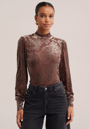 Femme portant un top ajusté en velours marron à motif floral, manches longues et col haut, associé à un jean noir taille haute et des boucles d'oreilles créoles dorées.