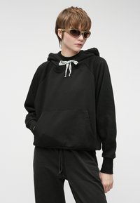 Μαύρη κουκούλα φούτερ με τσέπη μπροστά και κορδόνια. Διαθέτει ραφές ράγκλαν και ριμπ μανσέτες. Φοριέται με ταιριαστά σκούρα παντελόνια.