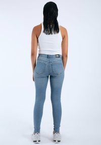 Jeans ajustados de color azul claro con bolsillos traseros, cintura alta y una etiqueta negra en la cinturilla, combinados con una camiseta sin mangas blanca y zapatillas blancas.