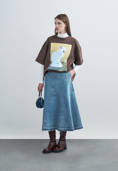Καφέ φαρδιά μπλούζα με γραφικό λευκής γάτας, συνδυασμένη με μπλε τζιν μακριά φούστα και γυαλιστερά μπορντό μποτάκια μέχρι το γόνατο.