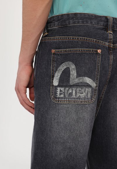 I jeans di denim scuro presentano una tasca posteriore decorativa con cuciture e un design del logo grigio. Gli accessori includono rivetti in rame e cuciture a contrasto.