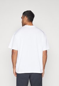 Hvid bomulds T-shirt med løs pasform, korte ærmer og rund halsudskæring, vist fra ryggen på en neutral baggrund.