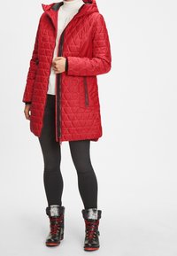 Rød quiltet frakke med hætte, med lynlås og sidelommer. Sættes sammen med sorte bukser og sorte støvler med pelskant og røde snørebånd.