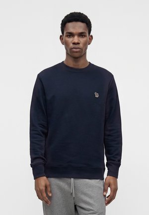 Ung sort mand iført en marineblå sweatshirt med et lille farverigt zebraemblem på brystet og grå joggingbukser, stående mod en ensfarvet baggrund.