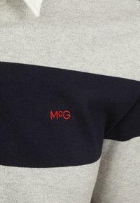 Gebreide trui met grijs en marineblauwe strepen; heeft een rood "McG" logo. De stof heeft een zachte textuur en is lichtgewicht.